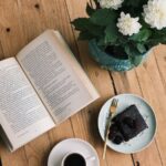 Charming scene of an open book, coffee, and cake beside flowers on a rustic wooden table.